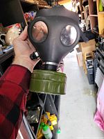 Front view of black rubber gas mask with two round eye lenses and attached green cylindrical filter canister held in hand.