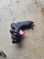 Photo of black electric hot glue gun with red trigger and coiled power cord on concrete floor.