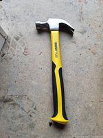 Photo showing a claw hammer with a yellow and black handle on a rough concrete surface.