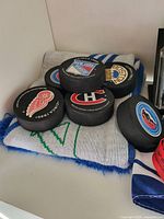 Photo showing five black hockey pucks featuring NHL team logos including New York Rangers, Boston Bruins, Detroit Red Wings, Montreal Canadiens, and a mascot puck on a knit scarf.