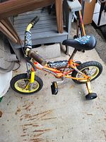 Kids bike with training wheels, predominantly orange with yellow front fork, black seat, and brown grips, shown on concrete floor outside.