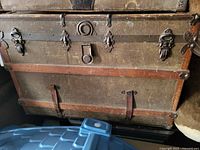 Front view of large vintage trunk showing woven material, wooden slats, leather straps, metal locks and circular metal ring detail.
