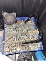 Blue compartment tray filled with a variety of metal screws of different sizes. Includes a black curved hand tool on top and a round speedometer gauge to the left. Bottom right shows part of a blue handheld power tool.