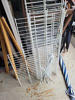 Photo of multiple white metal wire shelving racks and support bars leaning against a wall on a concrete floor.