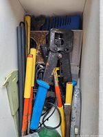Box with miscellaneous hand tools including pliers, screwdrivers, and a crimping tool with colored handles, some showing rust and wear.