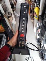 Side view of black NOMA surge protector showing 12 outlets, including a highlighted red outlet, and USB ports at the bottom. Surge protection indicator lights and labels visible.