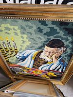 Photo of framed needlepoint artwork showing a seated elderly man in prayer shawl reading with menorah candles lit beside him.