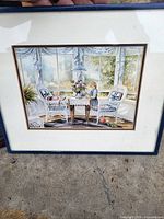 Framed print showing a child standing by a table with a doll sitting in a chair in a sunlit room with white wicker furniture and floral decor.