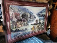 Framed landscape painting showing a wooden cabin near a serene lake surrounded by mountains and trees.