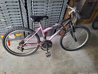 Photo shows a pink and black mountain bike leaning against a stack of plastic storage containers. The bike has front suspension forks, knobby tires, a padded seat, and multiple-speed gears with pedals included. There is a basket behind the seat on the right side of the bike.
