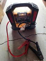 Front view of the black and yellow portable jump starter with red and black clamps lying in front. Handle on top is visible, as well as control panel and labels behind the plastic cover.