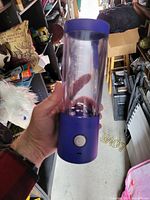 Hand-held purple portable blender with clear plastic container and white button on purple base, displayed among various household items.