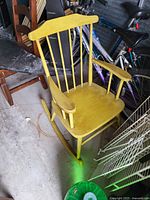 Yellow wooden rocking chair with vertical slat backrest, armrests, and rockers, shown in a garage or storage area.