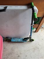 Photo of a children's kick scooter with green foam grips on the handlebars, green wheels, and a blue deck with text on it. The scooter is folded and resting against a white table.