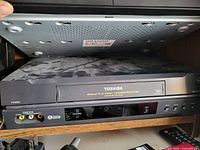 Front view of black Toshiba VHS player model with V-Audio Hi-Fi, control panel, input ports and buttons, placed on wooden shelf.