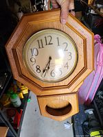 Octagonal wooden wall clock held by hand, showing clock face with numbers 1 to 12 and hour, minute, and second hands. Pendulum window at the bottom of the clock body is visible.