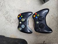 Two black Xbox 360 wireless controllers on concrete surface with visible buttons, thumbsticks, and directional pads.