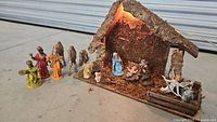 Full view of the nativity scene set arranged outdoors showing stable with bark roof, moss interior, and figurines: Mary, Joseph, baby Jesus in manger, three wise men, angel, camels, and sheep. The set is lit from inside producing a warm glow.
