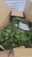 Packed artificial Christmas tree with green foliage inside an opened box showing tree parts and instruction paper.