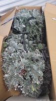 Artificial Christmas tree folded inside a damaged box, showing snowy blue-green pine needles and plastic stand parts.