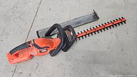 Full view of the Black & Decker cordless hedge trimmer lying on the floor, showing the orange and black casing with the attached 22-inch blade.