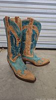 Photo showing pair of turquoise and brown leather cowboy boots with eagle and heart embroidery design, medium heel, and pull tabs, positioned side by side.
