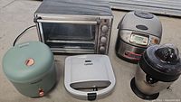 Photo showing the lot of four kitchen appliances including Cuisinart toaster oven (stainless steel), Black & Decker rice cooker (silver and black), Chaceef green rice cooker, sandwich maker in white, and black juicer.