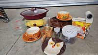 Overall lot view showing various kitchenware items including 5-quart Crock-Pot, ceramic owl dishes, measuring cup, wooden tray, food chopper, and can opener.
