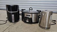 Photo showing the three kitchen appliances together: air fryer, slow cooker, and electric kettle on concrete floor