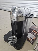 Full view of Nespresso pod coffee machine, black with chrome top and side water tank. Machine shows minor surface marks. Box of Aieve refillable coffee capsule kit is beside it.