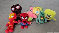 Photo showing full set of six plush toys including two Spider-Man variants (classic and black suit), Iron Man, Patrick Star, Plankton, and SpongeBob SquarePants arranged closely on concrete floor.