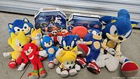 Overview of the full lot showing 2 RC cars in original packaging and 12 Sonic the Hedgehog plush toys including Sonic, Tails, Knuckles, and Dr. Eggman characters arranged together