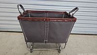 Front view of laundry cart showing gray canvas body with brown trim, metal frame base, and caster wheels. Canvas handles are visible.