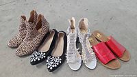 Four pairs of shoes: leopard print ankle boots, black flats with white bead embellishments, silver strappy heels, and red slide sandals on concrete floor.
