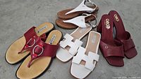 Four pairs of sandals and slides arranged on floor: red heeled slides, white H-strap slides, white metallic ankle strap thong sandals, red thong sandals with metal rings.