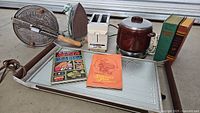 Photo showing the Detonator popcorn popper, vintage electric iron, white electric two-slot toaster, brown ceramic slow cooker with lid, three cookbooks, and electric cooking tray.