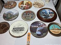 Top view photo showing eleven collectible plates laid flat with various scenic and historical imagery, one silver plated spoon in packaging, and three black metal plate stands.