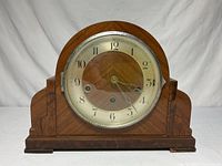 Front view of wooden antique mantle clock with round dial, glass cover closed.