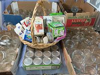 Overview of multiple glass jars in boxes and basket with unopened boxes of jelly jars, box of lids, and assorted bands and lids.