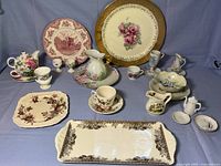 Full view of the mixed china pieces showing various plates, pitchers, cups, and servingware with floral and scenic designs. The large plate with red pattern shows a taped chip.