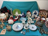 Wide shot showing entire Christmas decoration set including plates, mugs, snowman figurines, Santa, and other ceramics on teal linen.