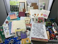 Wide view of assorted craft papers, envelopes, coloring sheets with cartoon and floral designs, stamps, scissors, tape, adhesives spread out on a surface.