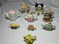 Eight decorative teapots and figurines displayed on gray fabric, showing various shapes and colors including houses, cabbage, and elephant figurine.