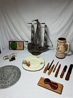 Full lot on table including metal sailboat sculpture, ceramic stein, small model T cars, decorative plate, set of wood-handled knives, knife rack, and coin plaque.