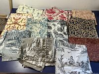 Several pieces of vintage fabrics with toile de Jouy and floral patterns laid out on a blue table, showing various colors and designs such as cream, red, blue, beige, burgundy, and black prints