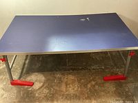 Front view of the folding craft table showing full blue surface and metal legs with red plastic feet and knobs