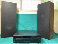 JVC multi disc CD player XL-F108BK positioned between two vintage tall floor speakers with black fabric covers.