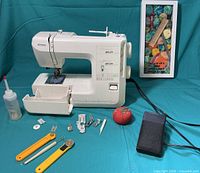 Full view of Kenmore sewing machine with attached bobbin cover, various sewing accessories laid out in front, small oil bottle, and yarn wall art in the background.