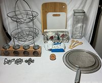 Photo of all kitchen items including two-tier metal fruit basket, wooden cutting board, tall glass jar, set of small jars in a wooden holder, ceramic tea set, metal splatter screen, wooden spoons, metal cookie cutters, small ceramic lid, and box of tissues all arranged on a white surface.