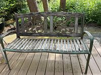 Front view of rustic garden bench with wood slat seat, green metal frame, and decorative floral backrest, showing wear and weathering.